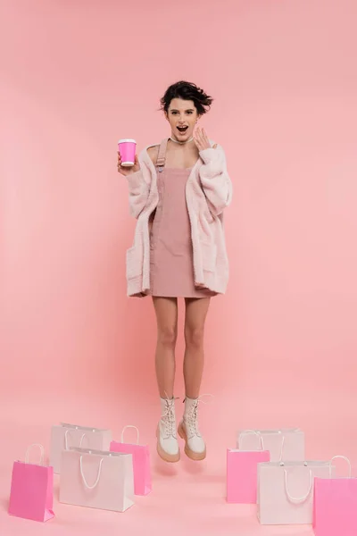 Longitud completa de la mujer asombrada con café para ir levitando cerca de bolsas de compras sobre fondo rosa - foto de stock