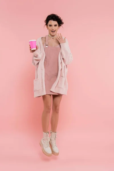 Pleine longueur de femme excitée en robe de sangle et confortable cardigan lévitant avec tasse en papier sur fond rose — Photo de stock