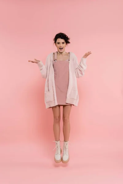 Full length of excited woman in strap dress and fluffy cardigan levitating on pink background — Stock Photo