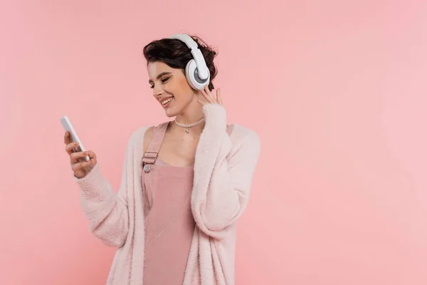 Щаслива брюнетка жінка в бездротових навушниках дивиться на мобільний телефон ізольовано на рожевому — стокове фото