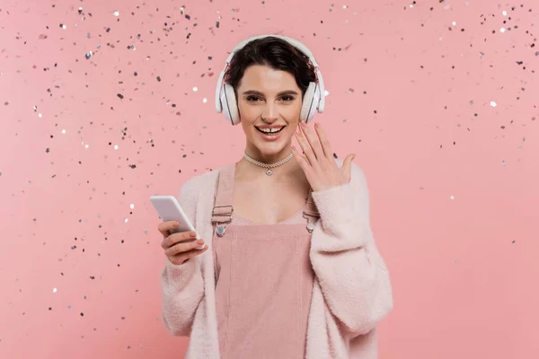 Cheerful brunette woman in wireless headphones holding smartphone under confetti on pink background — Stock Photo