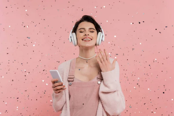 Mulher alegre com olhos fechados segurando celular e ouvindo música em fones de ouvido sem fio perto de confete em rosa — Fotografia de Stock