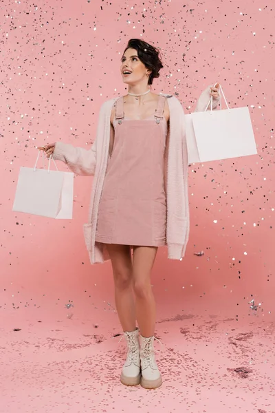 Pleine longueur de femme heureuse et à la mode debout avec des sacs à provisions sous confettis sur rose — Stock Photo