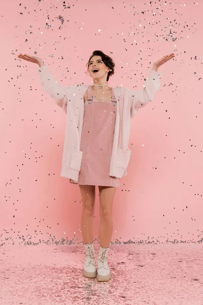 Comprimento total da mulher alegre em cardigan fofo pegando confete com as mãos levantadas em rosa — Fotografia de Stock