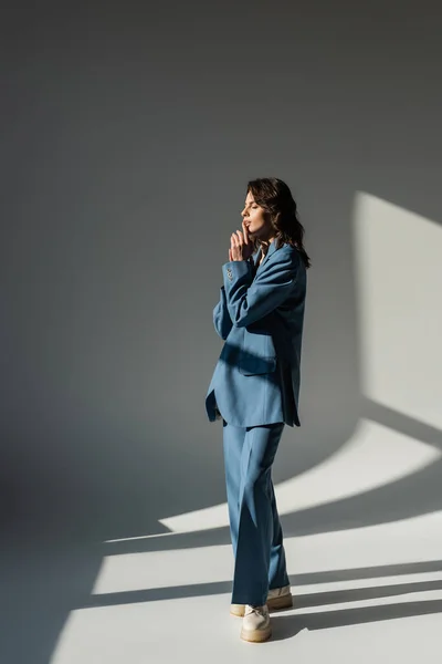Longitud completa de la mujer de moda en traje azul de pie con los ojos cerrados sobre fondo gris con iluminación - foto de stock