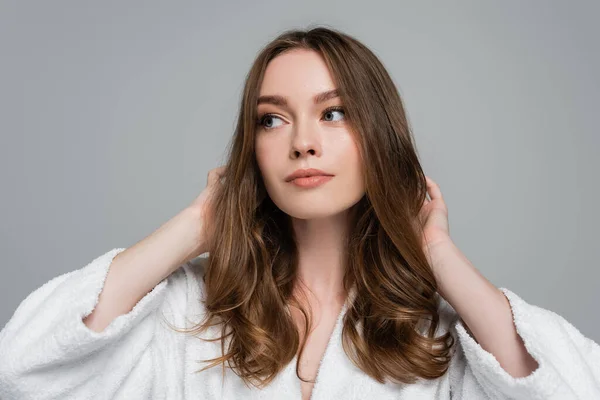 Young woman in bathrobe touching shiny and healthy hair while looking away isolated on grey — Stock Photo