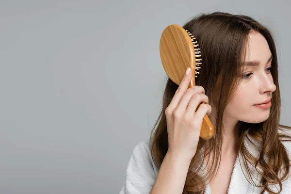 Bonita jovem mulher em roupão escovando cabelo brilhante e saudável isolado em cinza — Fotografia de Stock