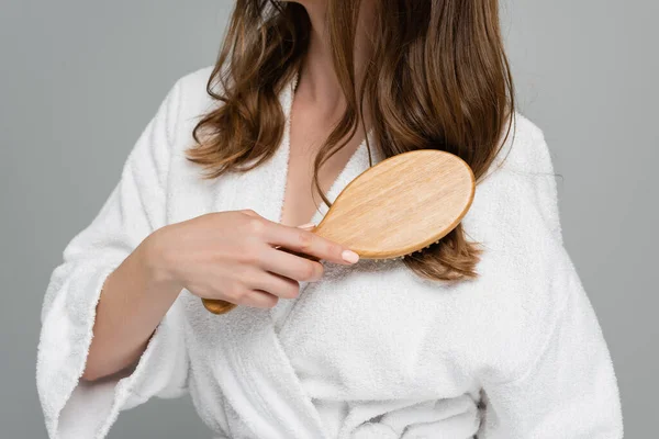 Vista cortada de jovem mulher em roupão de banho branco escovando cabelo brilhante e saudável isolado em cinza — Fotografia de Stock
