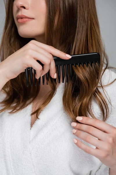 Vista cortada de mulher jovem escovando cabelo brilhante e saudável com pente de plástico isolado em cinza — Fotografia de Stock