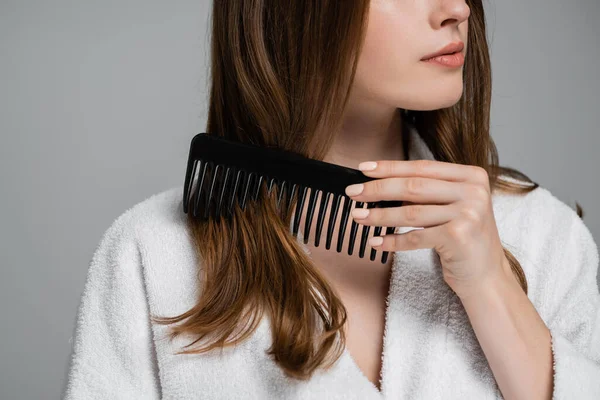 Teilansicht einer jungen Frau, die glänzendes und gesundes Haar mit einem auf grau isolierten Kunststoffkamm bürstet — Stockfoto