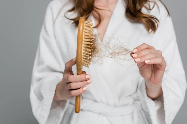 Обрезанный вид молодой женщины, держащей деревянную щетку для волос и вытягивающей поврежденные волосы, изолированные на сером — стоковое фото