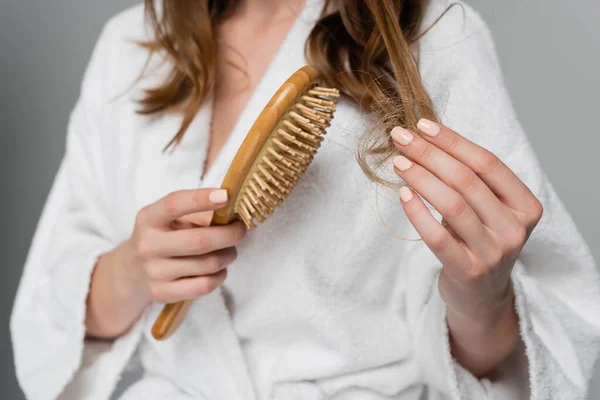 Visão parcial da jovem segurando escova de cabelo de madeira enquanto puxa o cabelo danificado isolado no cinza — Fotografia de Stock