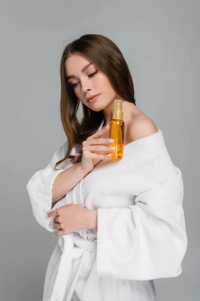 Брюнетка молодая женщина с блестящими волосами держа бутылку с маслом изолированы на сером — стоковое фото