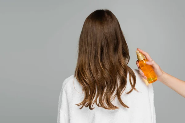 Профессиональный стилист, держащий бутылку с маслом рядом с молодой женщиной с блестящими волосами, изолированными на сером — стоковое фото