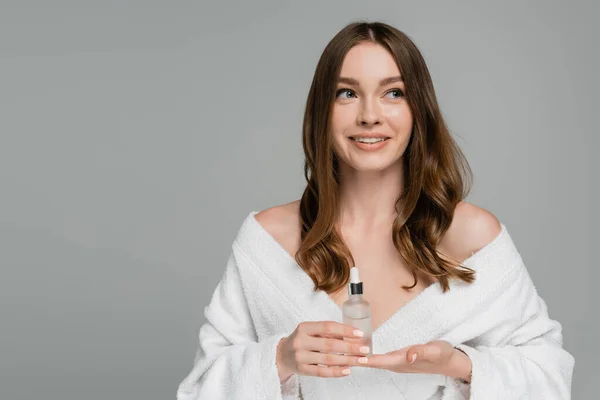 Happy young woman with shiny hair holding bottle with serum isolated on grey — Stock Photo