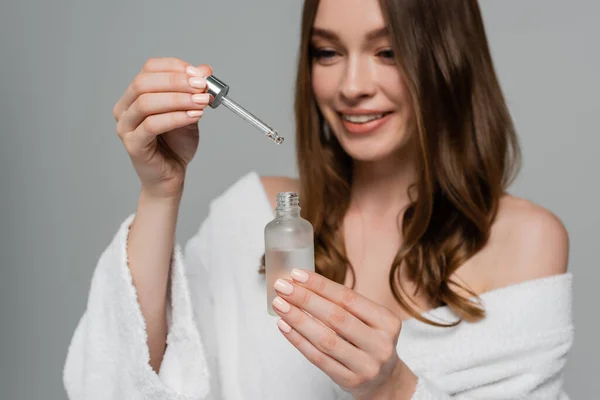 Cheerful young woman with shiny hair holding bottle and pipette with serum isolated on grey — Stock Photo