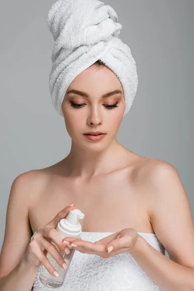 Young woman with bare shoulders and towel on head holding bottle with cleansing foam isolated on grey — Stock Photo