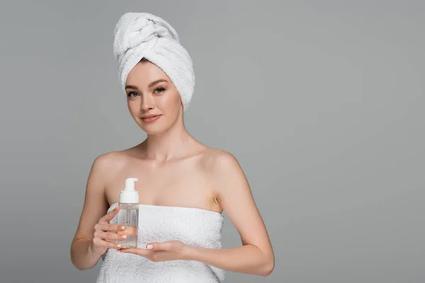 Giovane donna sorridente con spalle nude e asciugamano sulla testa bottiglia con schiuma detergente isolata su grigio — Foto stock