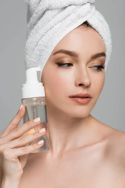 Portrait of young woman with towel on head holding bottle with cleansing foam isolated on grey — Stock Photo