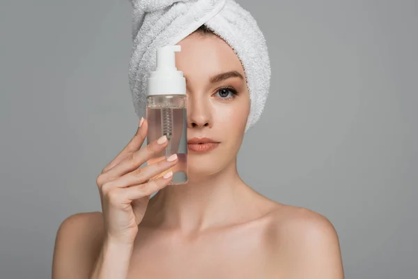 Junge Frau mit nackten Schultern und Handtuch auf dem Kopf hält Flasche mit Reinigungsschaum in Gesichtsnähe isoliert auf grau — Stockfoto