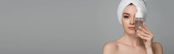 Young woman with bare shoulders and towel on head holding bottle with cleansing foam near face isolated on grey — Stock Photo