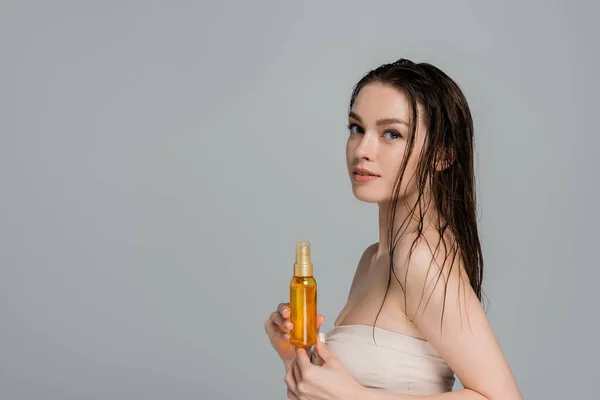Jolie jeune femme aux cheveux mouillés et aux épaules nues tenant bouteille avec de l'huile isolée sur gris — Photo de stock