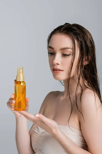 Jolie jeune femme aux épaules nues regardant bouteille avec de l'huile capillaire isolée sur gris — Photo de stock