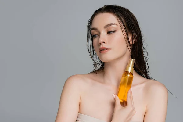 Jovem com ombros nus segurando garrafa com óleo de cabelo isolado em cinza — Fotografia de Stock