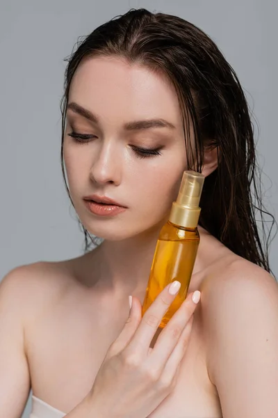 Портрет красивой молодой женщины с голыми плечами, держащей бутылку с волосяным маслом, изолированным на сером — стоковое фото