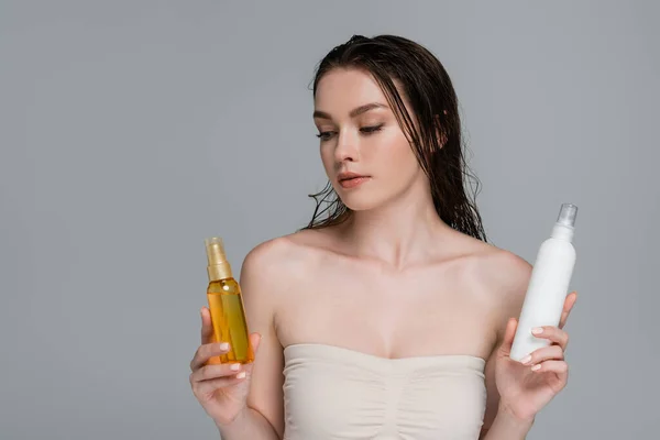 Hübsche junge Frau mit nassen Haaren und nackten Schultern, die zwischen Haarbehandlungsflaschen auf grau isoliert wählen — Stockfoto