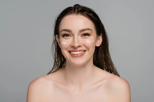 Joyeuse jeune femme aux cheveux mouillés et crème hydratante sur les joues isolées sur gris — Photo de stock