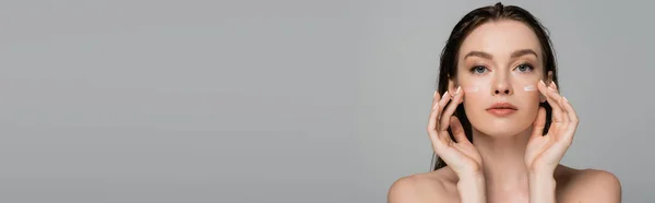 Giovane donna con capelli bagnati applicare crema viso sulle guance isolate su grigio, banner — Foto stock