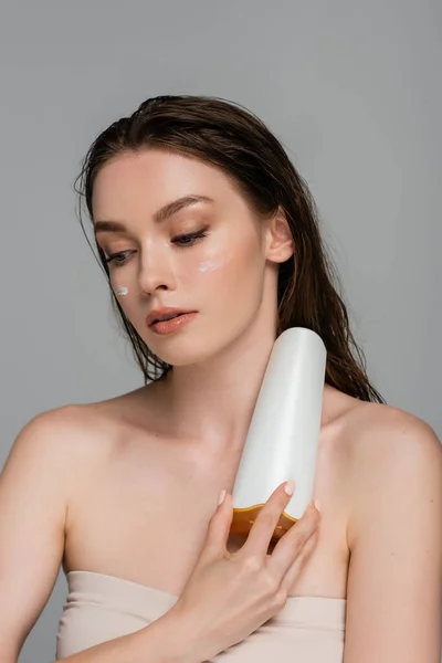 Pretty young woman with wet hair and moisturizing cream on cheeks holding bottle with shampoo isolated on grey — Stock Photo