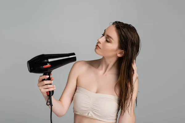 Красивая молодая женщина с мокрыми волосами с помощью фена, изолированного на сером — стоковое фото