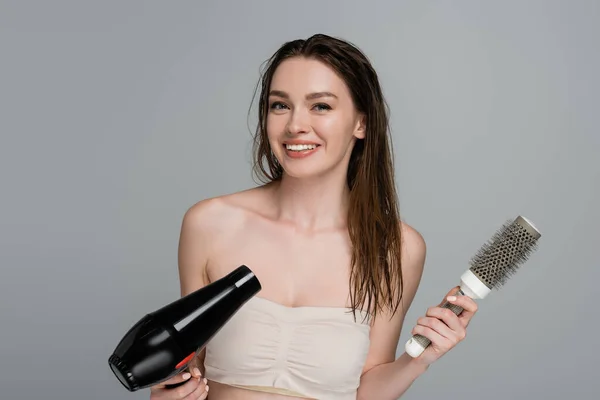 Cheerful young woman with wet hair holding round hair brush and hair dryer isolated on grey — Stock Photo