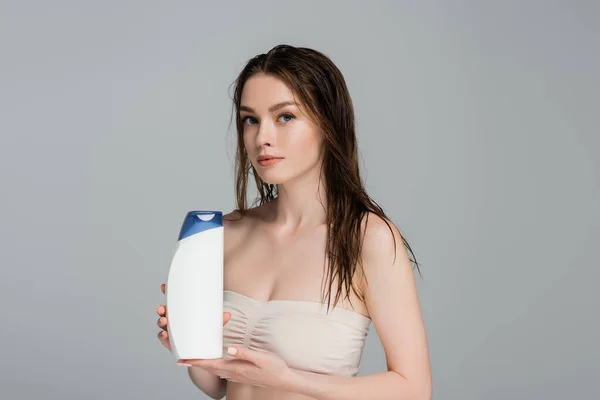 Гарна жінка з мокрим волоссям і голими плечима тримає пляшку шампуню ізольовано на сірому — стокове фото