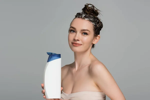 Cheerful young woman with wet foamy hair holding bottle with shampoo isolated on grey — Stock Photo