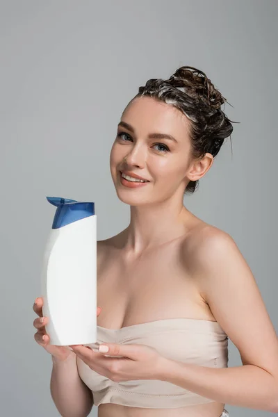 Smiling young woman with wet foamy hair holding bottle with shampoo isolated on grey — Stock Photo