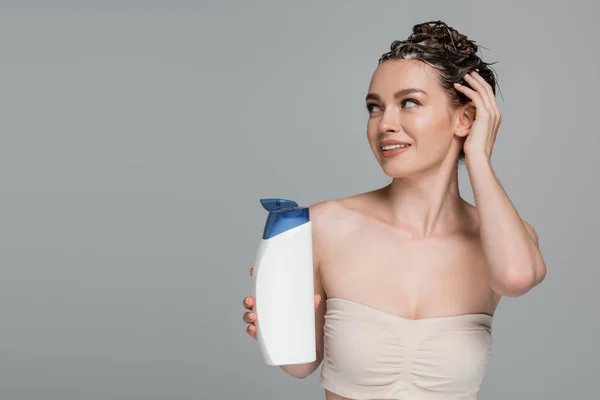Glückliche junge Frau mit nassen schäumenden Haaren, die Flasche mit Shampoo isoliert auf grau hält — Stockfoto