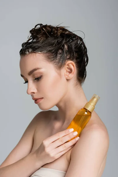 Brunette jeune femme aux cheveux mouillés tenant bouteille avec de l'huile de traitement isolé sur gris — Photo de stock