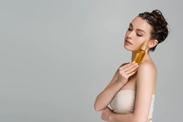 Brunette young woman with wet hair holding bottle with oil isolated on grey — Stock Photo