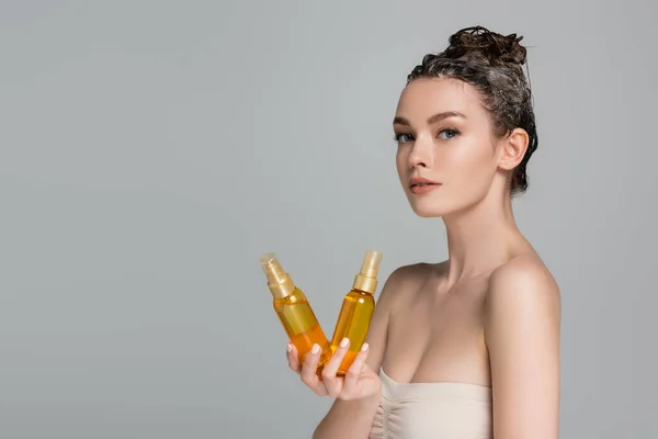 Brunette jeune femme aux cheveux mouillés tenant des bouteilles avec de l'huile de traitement isolé sur gris — Photo de stock