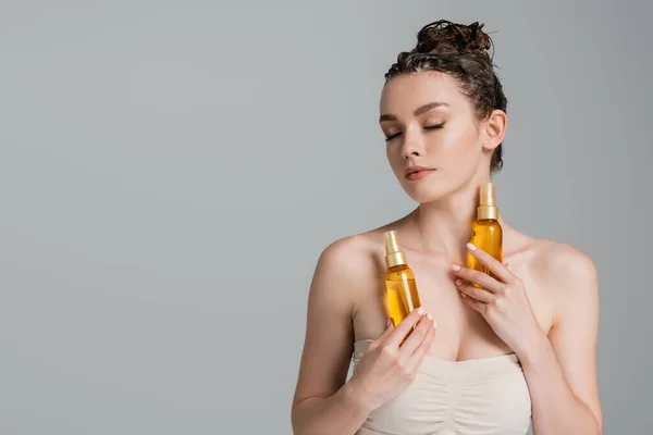 Brunetta giovane donna con gli occhi chiusi in possesso di bottiglie con olio per capelli trattamento isolato su grigio — Foto stock