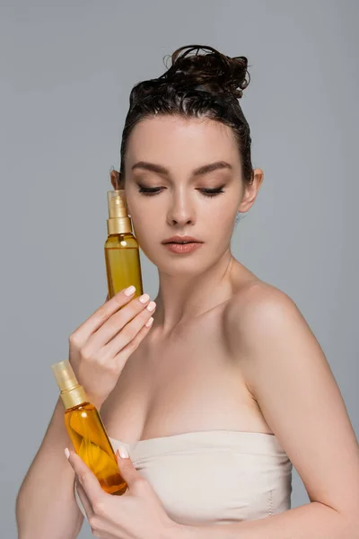 Jolie jeune femme aux cheveux mousseux mouillés tenant des bouteilles avec de l'huile de traitement isolé sur gris — Photo de stock