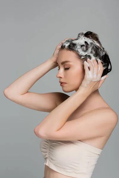 Молода жінка з закритими очима миє мокре і пінисте волосся ізольовано на сірому — стокове фото