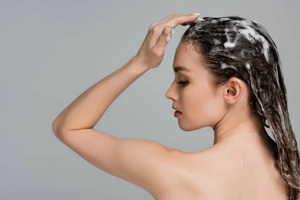 Vue latérale de la jeune femme aux yeux fermés lavant les cheveux mouillés et mousseux isolés sur gris — Photo de stock