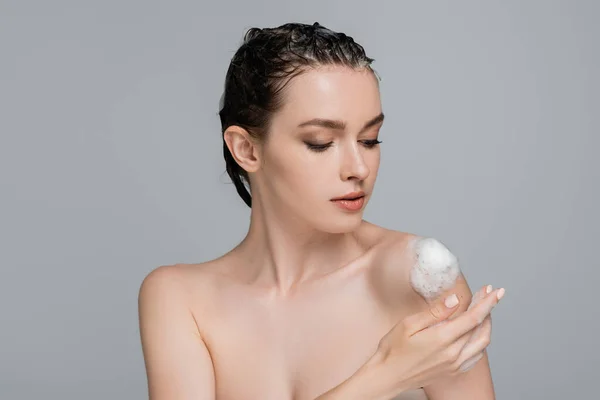 Jeune femme aux cheveux mouillés touchant épaule nue avec mousse blanche isolée sur gris — Photo de stock