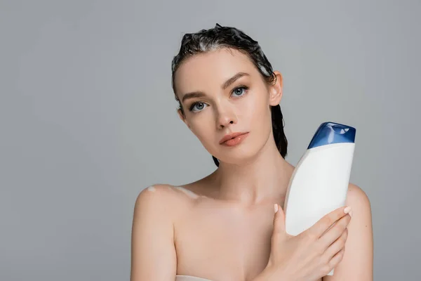 Brunette jeune femme aux cheveux mouillés tenant bouteille avec shampooing isolé sur gris — Photo de stock