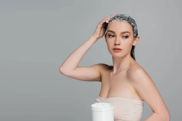 Jeune femme aux épaules nues appliquant un masque sur cheveux mouillés et tenant un bocal isolé sur gris — Photo de stock