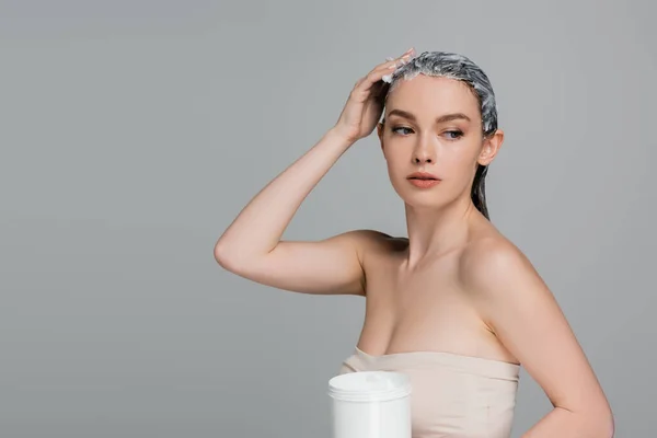 Young woman with bare shoulders applying mask on wet hair and holding jar isolated on grey — Stock Photo